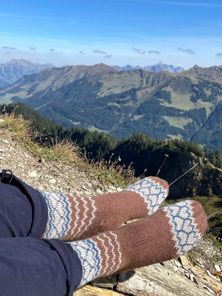 Braune Socke mit Hellbraunem und Blauen Einstrickmuster in Herzform vor einer Bergkulisse